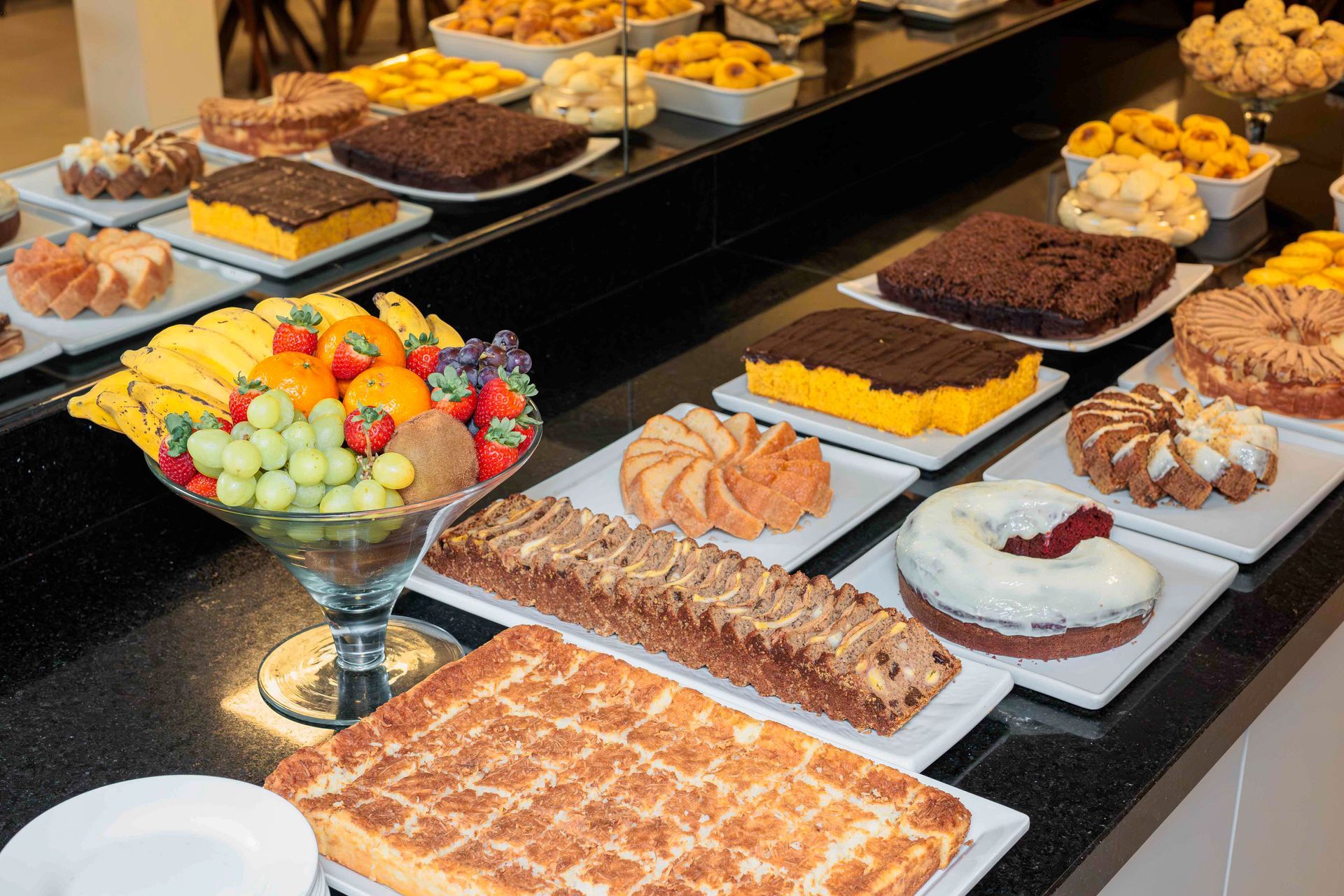 Mesa de buffet com taça de frutas (bananas, uvas, morangos, laranjas) e variedade de bolos.
