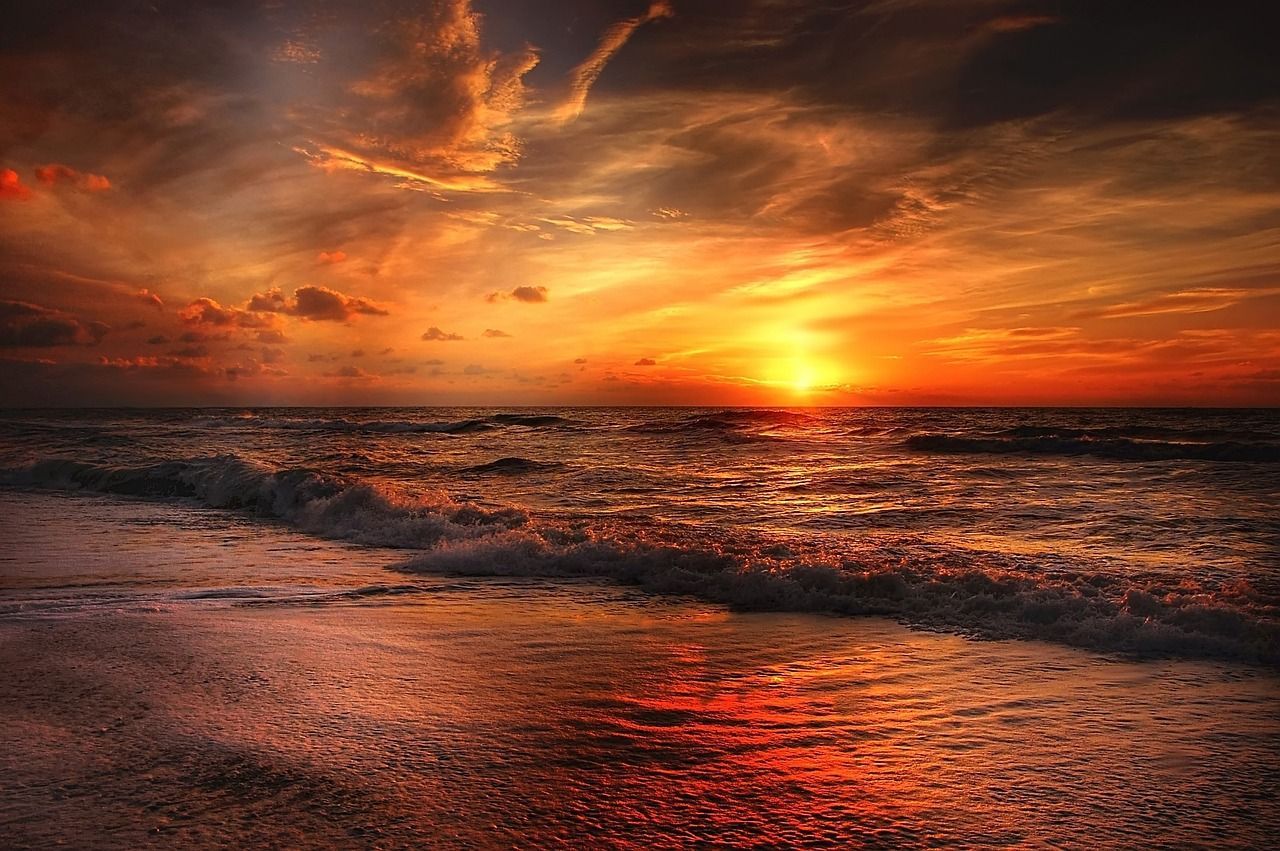 Pôr do sol no mar, céu alaranjado com nuvens e ondas suaves refletindo a luz na areia
