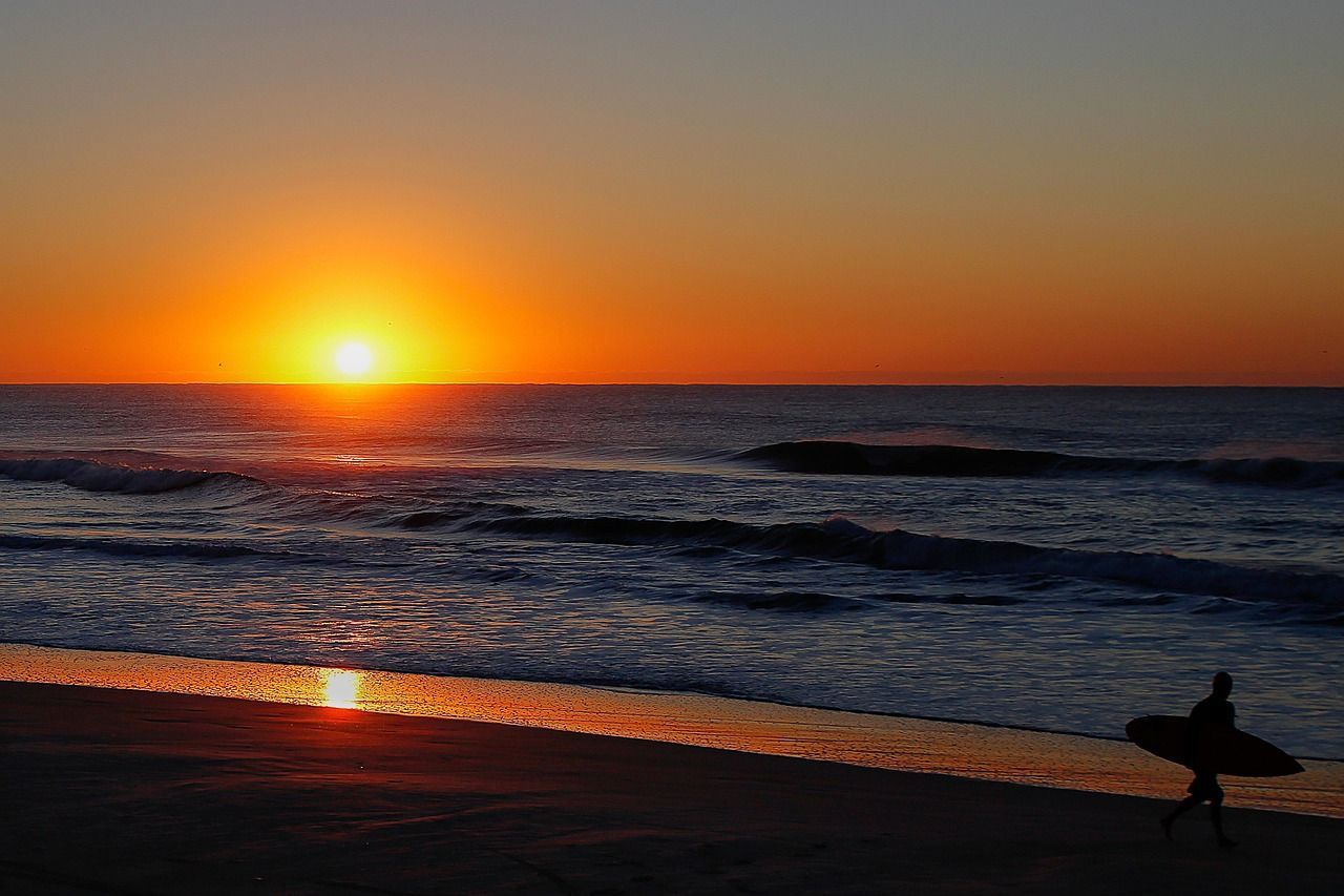Surfista caminhando na praia ao amanhecer com o sol nascendo no horizonte