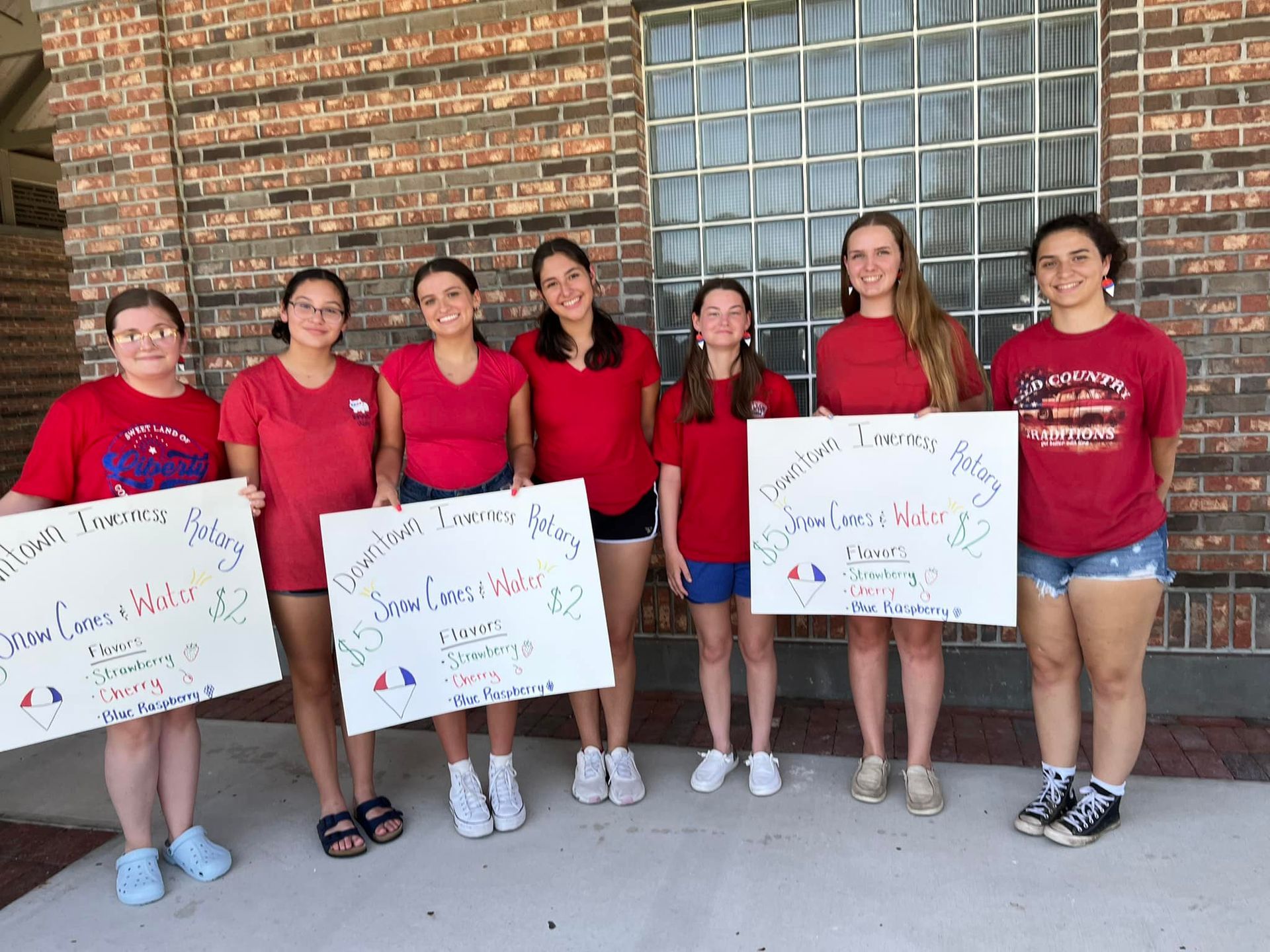A group of young women in red shirts are holding signs in front of a brick building.