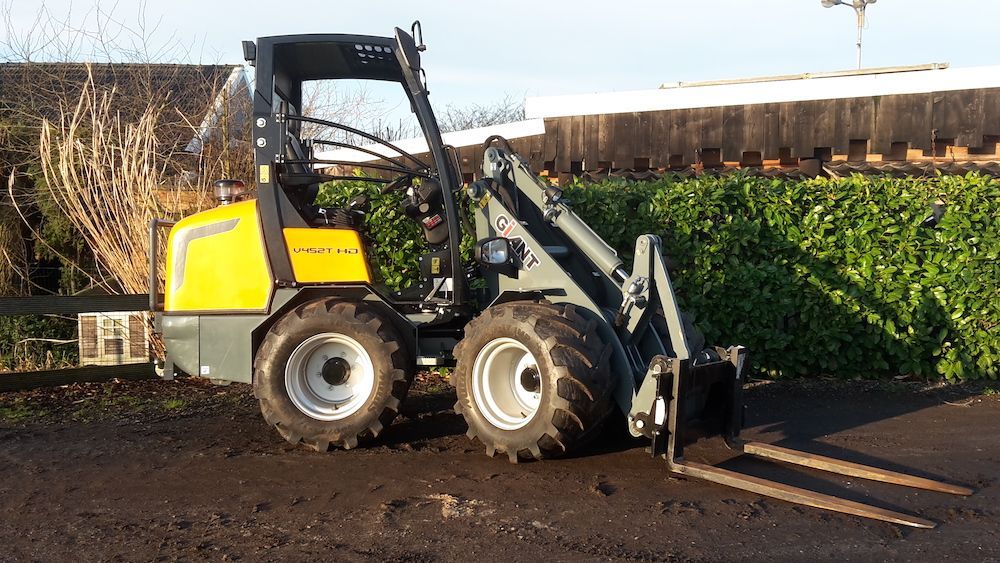 A yellow and gray forklift is parked in the dirt in front of a hedge.