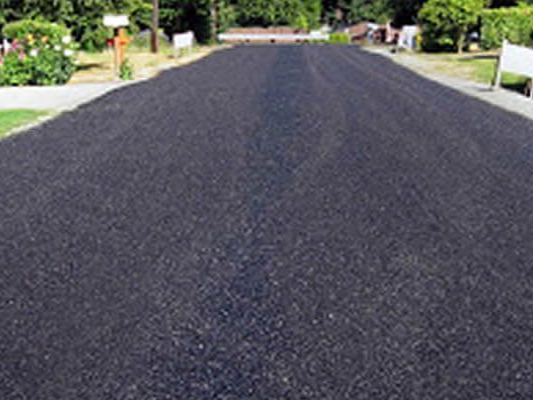 A freshly paved, black asphalt driveway stretches forward between green lawns and landscaping on a sunny day.
