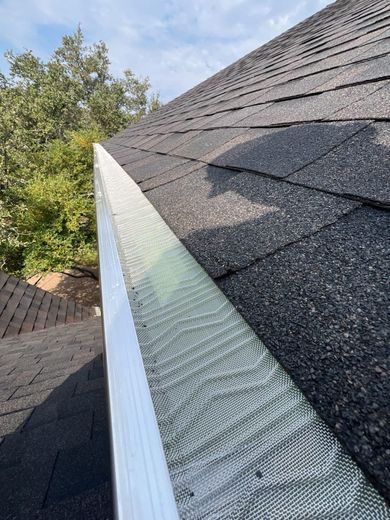 Close-up of a roof valley with dark shingles, white trim, and a metal rain diverter beside trees.