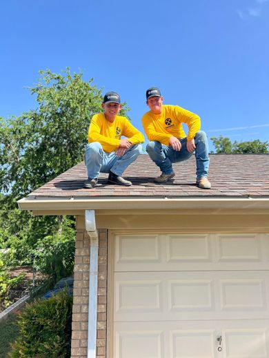 Two people in yellow shirts crouch on a garage roof under a clear blue sky.