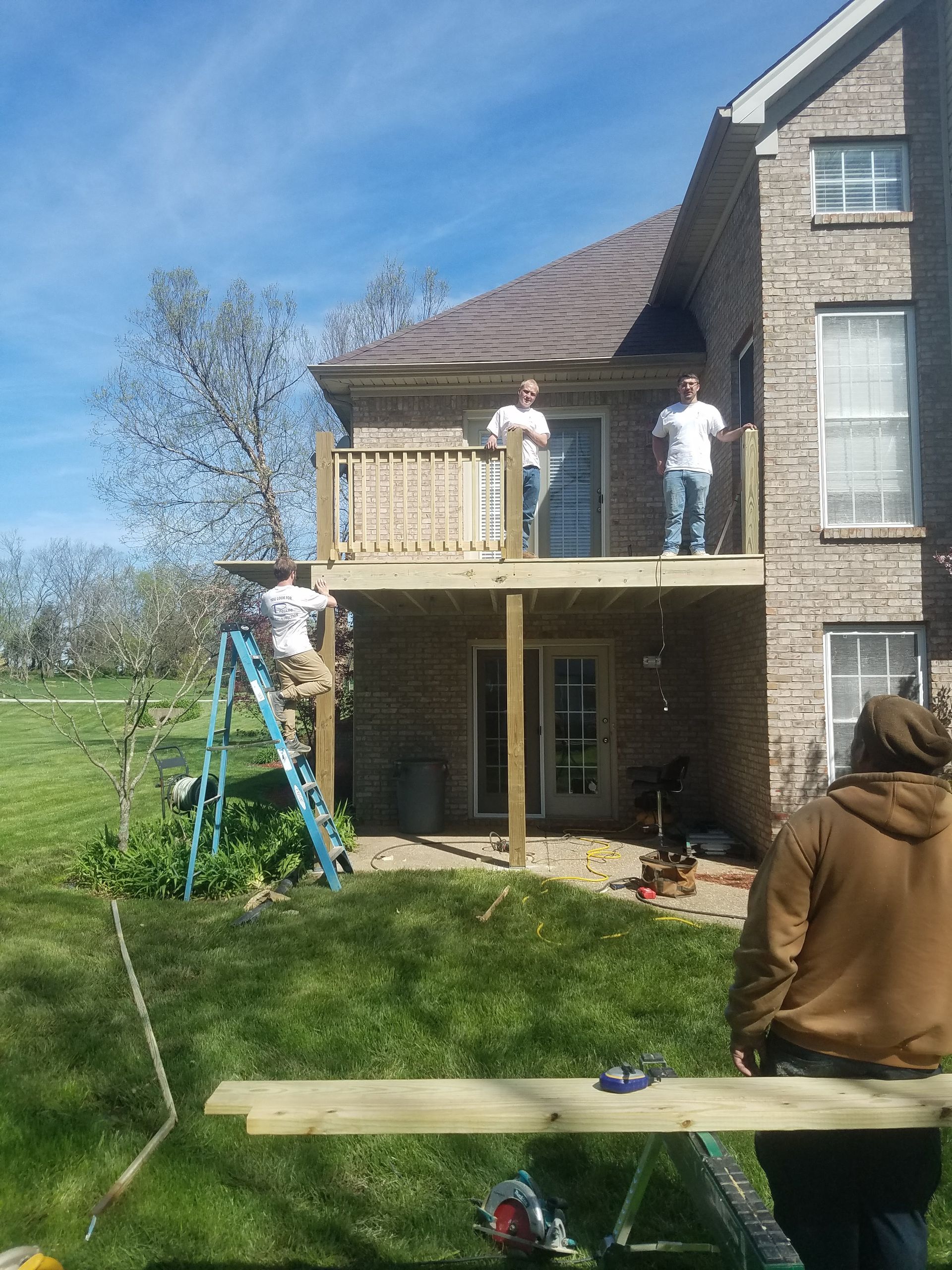 Two men are standing on a wooden deck in front of a brick house.