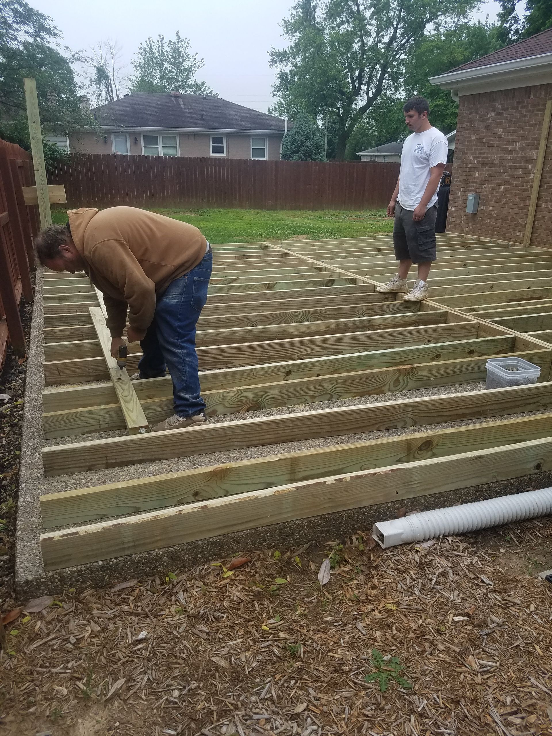 Two men are working on a wooden deck in a backyard.