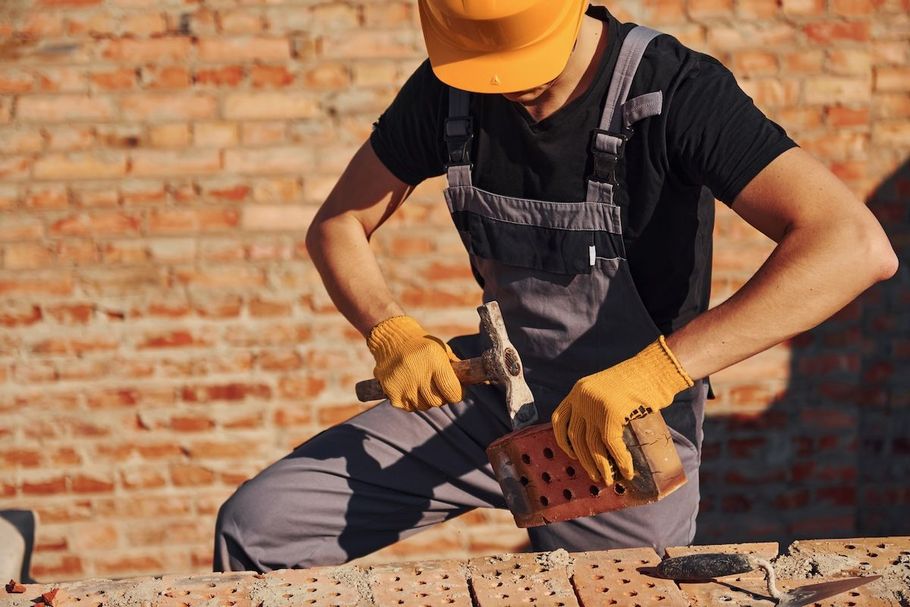 Construction worker in yellow hard hat and gloves using a tool on a brick wall.