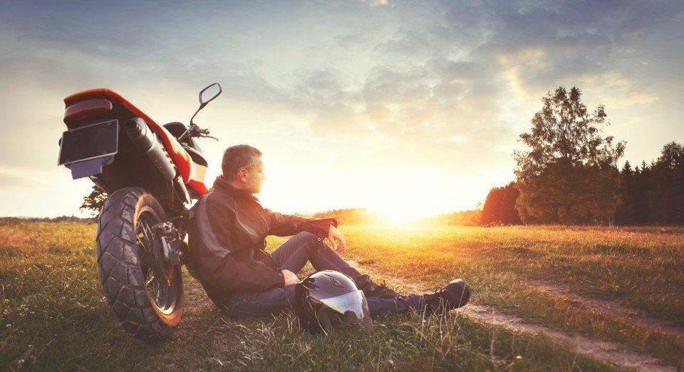Un uomo riposa appoggiato alla sua motocicletta in un campo al tramonto, con il casco a terra.