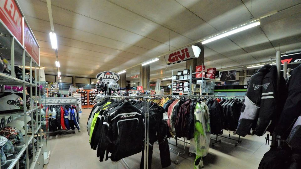 Interno del negozio di abbigliamento per motociclisti: espositori per vestiti con giacche, caschi su scaffali, luci sospese.