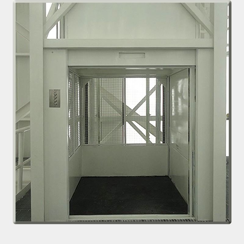 Pequeño ascensor industrial con paredes metálicas blancas y suelo oscuro, visto desde la puerta