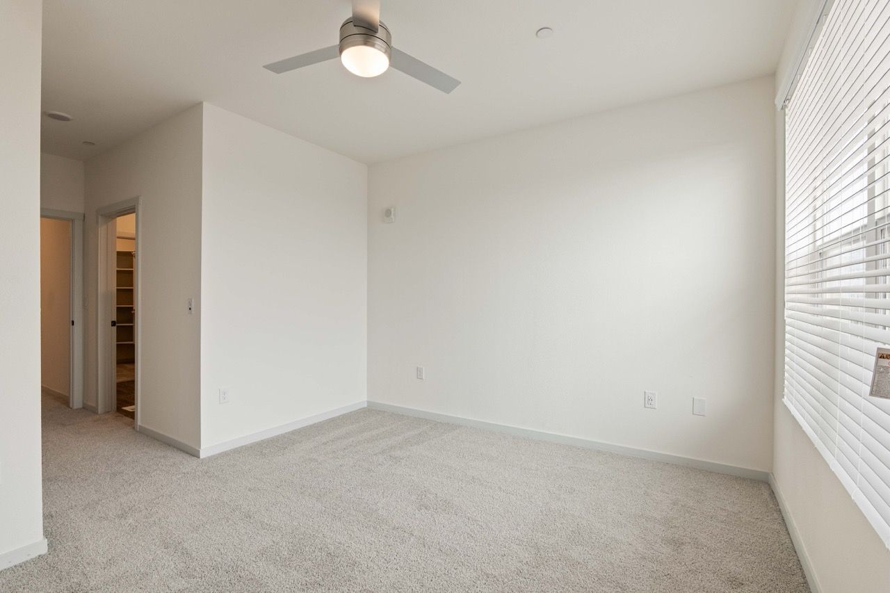 Empty apartment room with white walls, beige carpet, ceiling fan, and a large window with blinds.