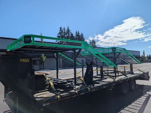 Green metal frame structure on a flatbed trailer, strapped down on a sunny day.