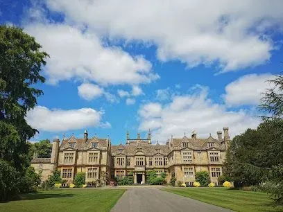 Large historic manor house with stone facade, green lawn, and blue sky with clouds