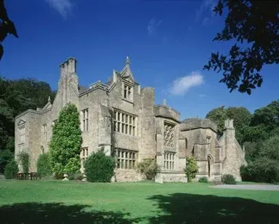 Large stone manor house with multiple gables and windows, set on a green lawn under a blue sky.