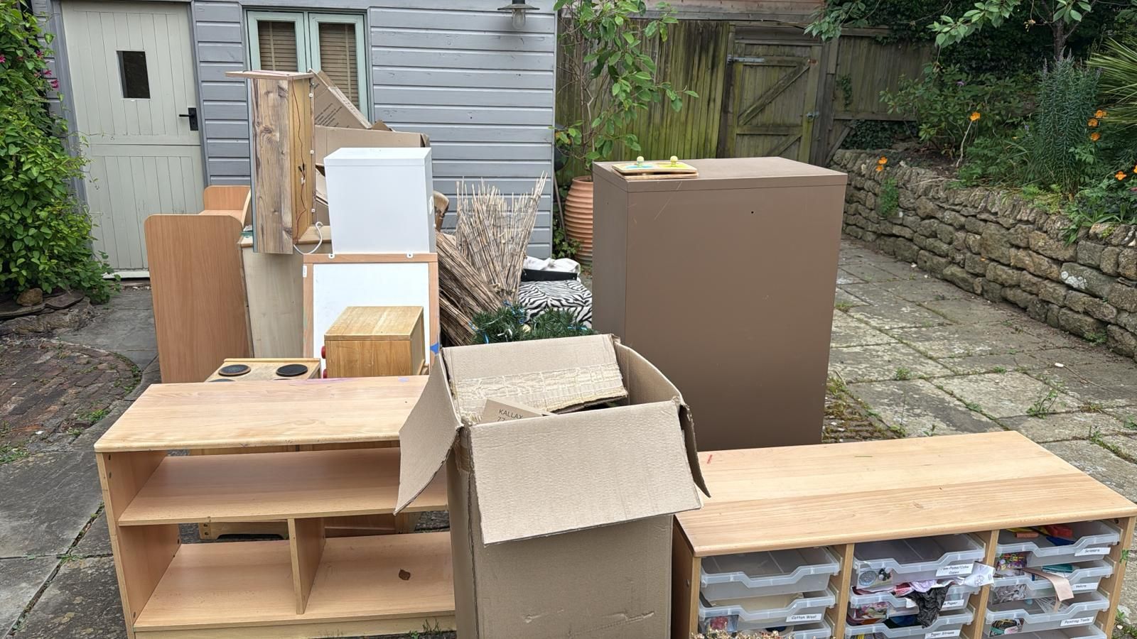 Staging area with cardboard boxes, wooden crates, and furniture on a driveway beside a house.