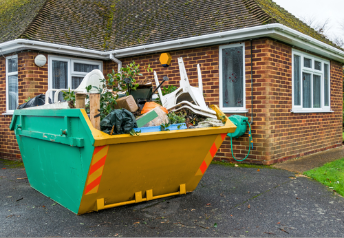 Green and yellow dumpster overflowing with trash in front of a brick house.