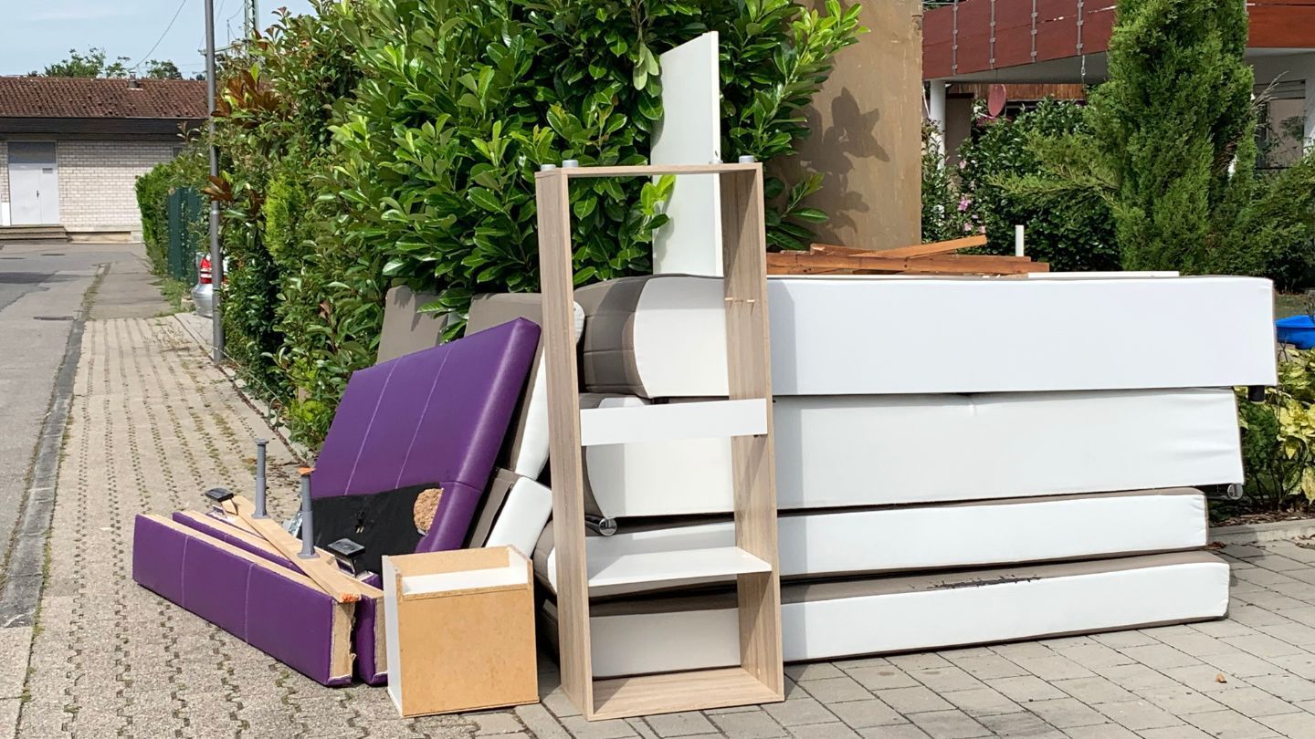 Discarded furniture on a sidewalk, including mattresses, shelving, and a purple headboard.
