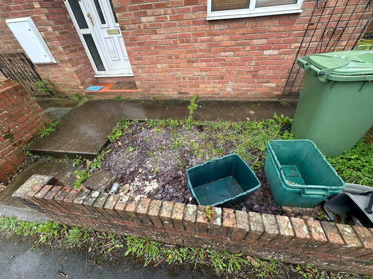 Front garden with muddy soil, two empty green bins, and a large green wheelie bin by a brick house.