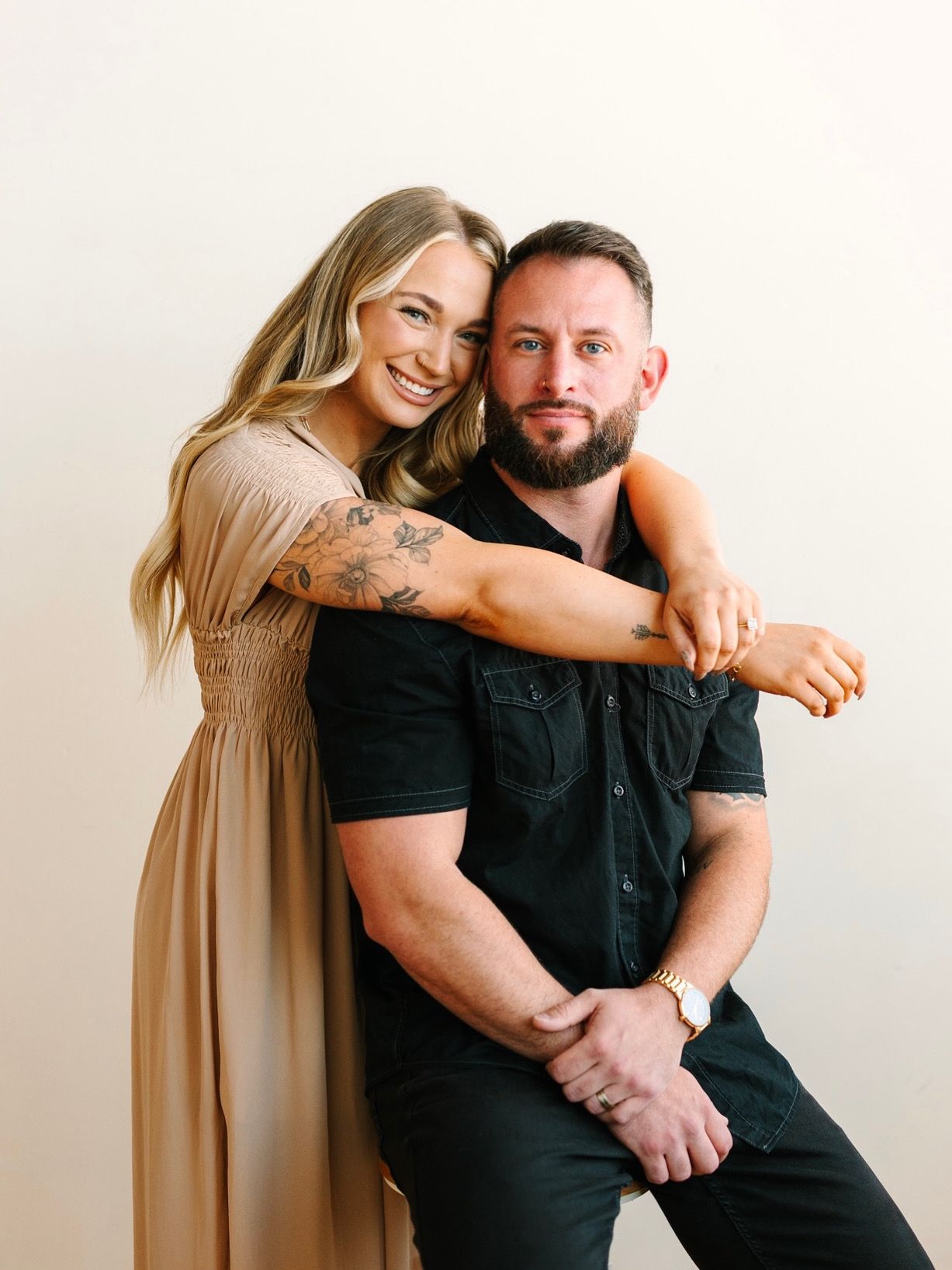 Woman in tan dress hugs man in black shirt. Both smile against a light background.