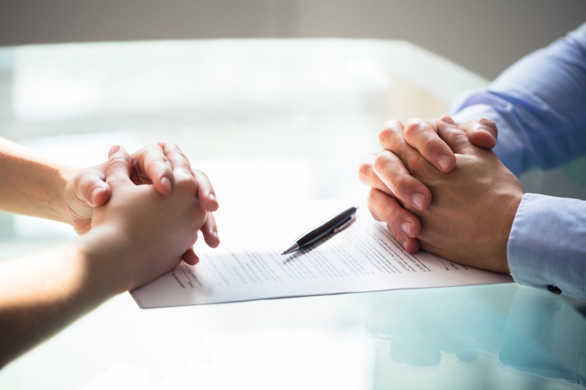 Two people sit at a glass table with their hands folded near a document and a pen, suggesting a professional meeting.