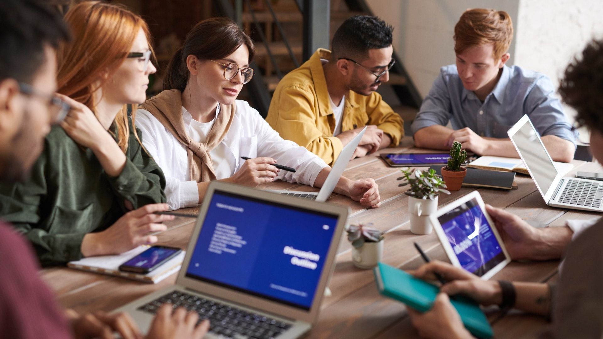 a group of people are sitting around a table with laptops and tablets .
