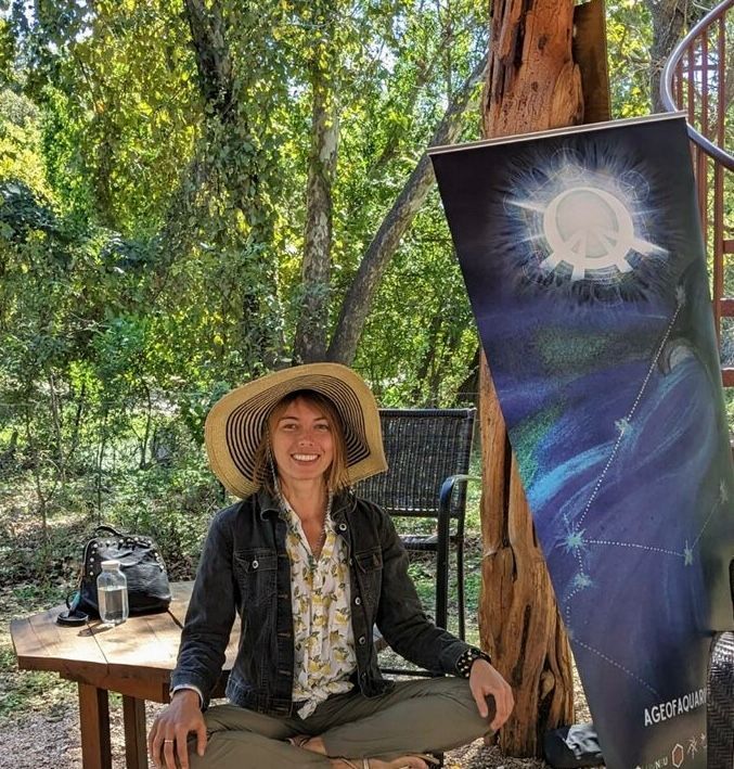 Woman in hat sits cross-legged outdoors, smiling. Dark jacket, patterned shirt. Next to poster of celestial art.