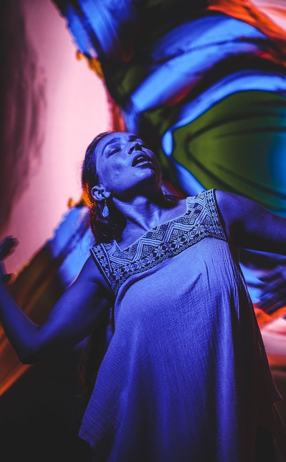 Native American Woman in traditional dress, arms raised, eyes closed, illuminated by colorful, abstract lighting.