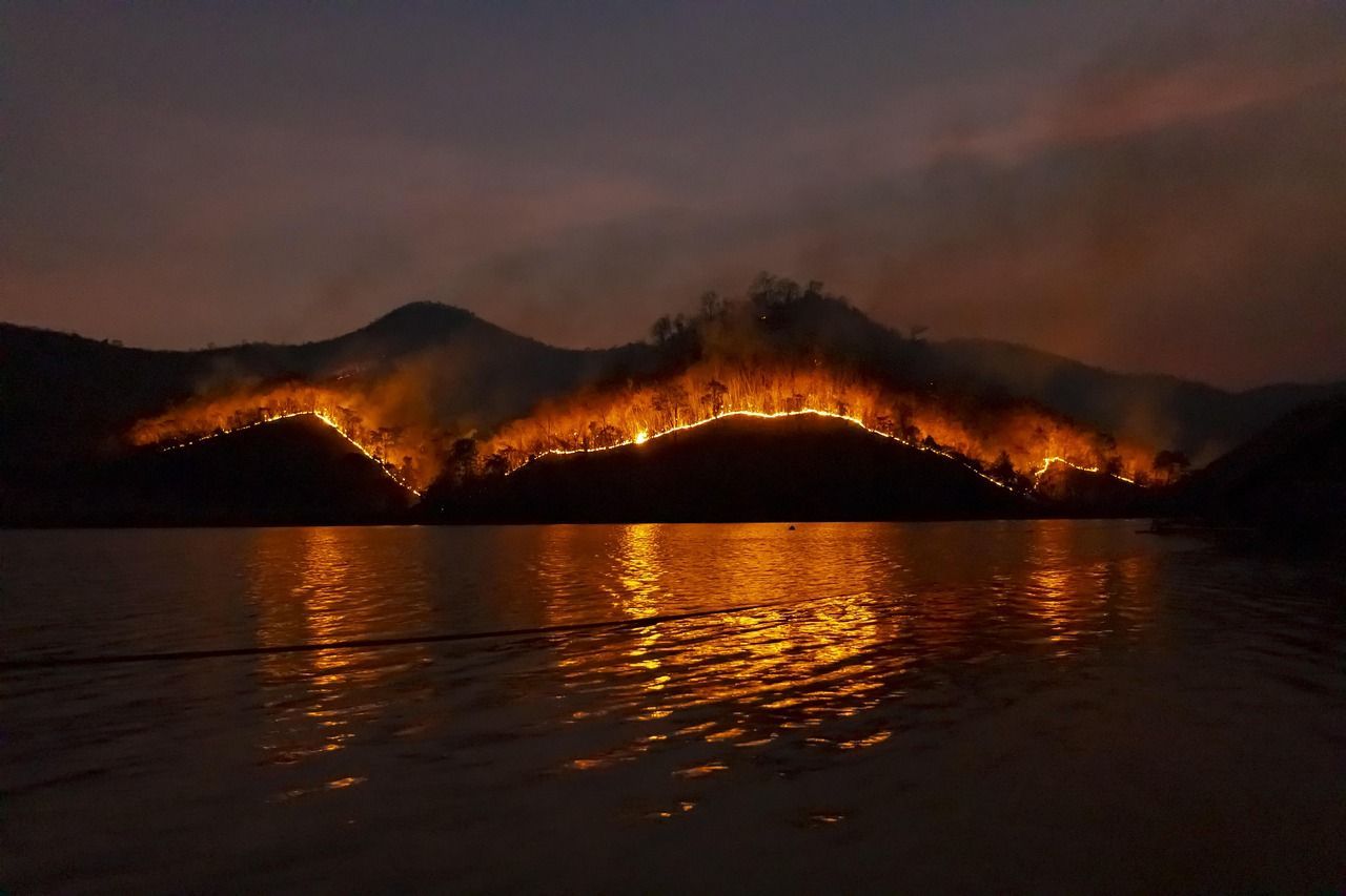 A nighttime view of a hillside ablaze, with flames reflected in the water below.