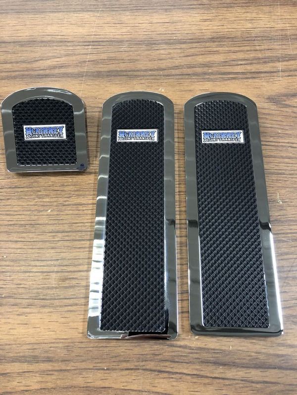 Three black and chrome car pedals with a textured center and a rectangular silver logo on a wood surface.
