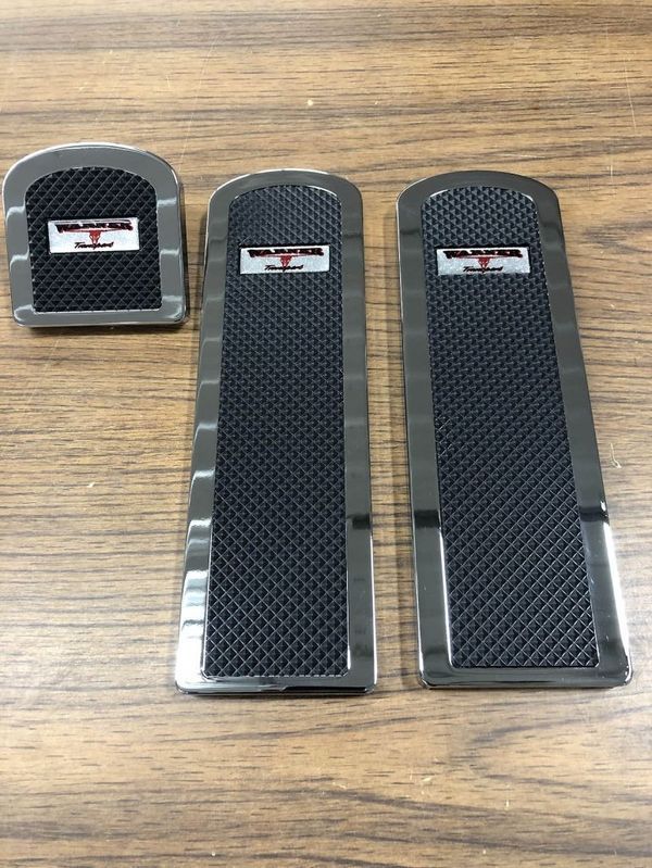 Three chrome and black car pedal covers on a wood surface, with a logo on each.