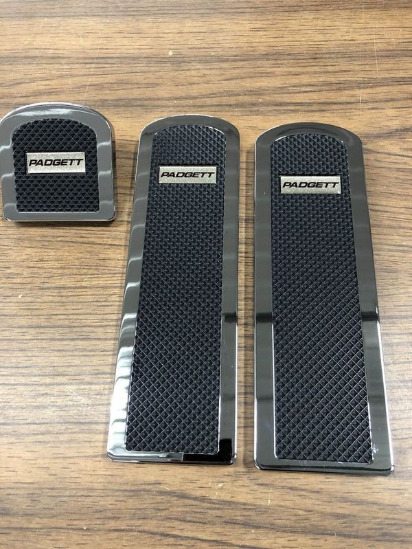 Set of three chrome and black Badgett brand car pedal covers on a wooden surface.