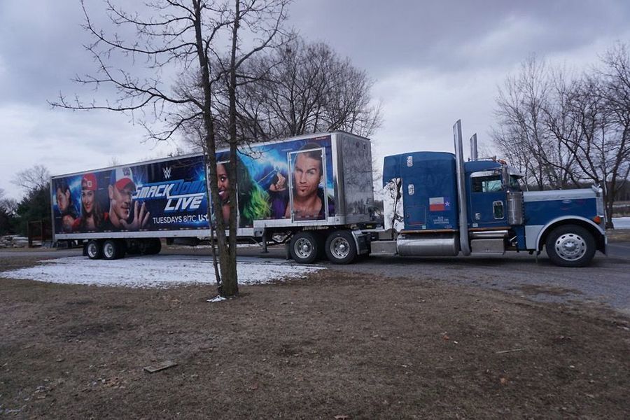 A semi-truck with a trailer wrapped in SmackDown Live graphics is parked outdoors.