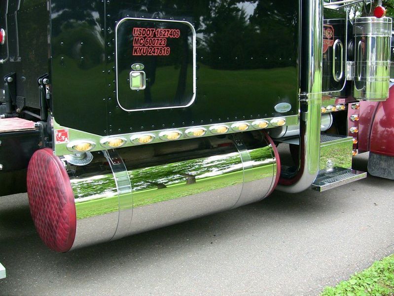 Chrome fuel tank and exhaust on a black semi-truck with red accents, parked on asphalt.