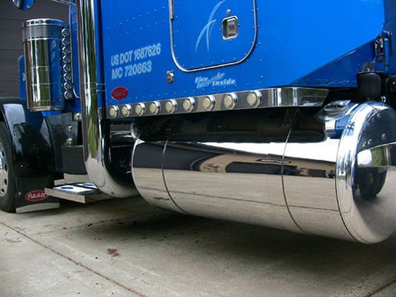 Blue semi-truck with shiny chrome fuel tank and exhaust, parked on concrete.