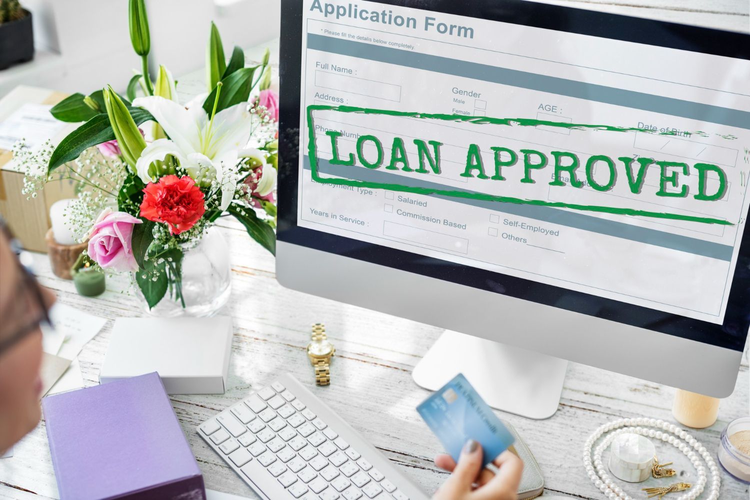 A woman is holding a credit card in front of a computer screen that says loan approved.