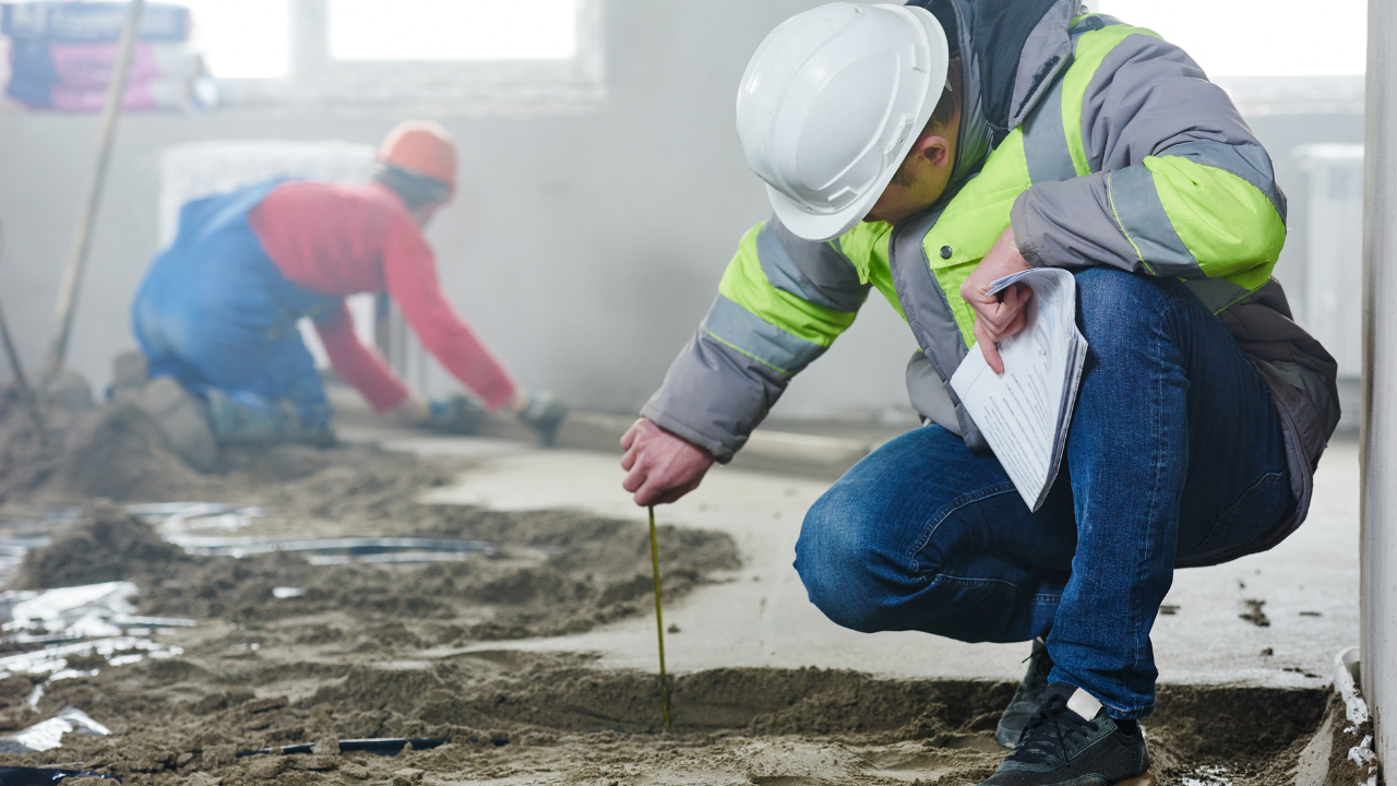 A worker in a white hard hat and high-visibility jacket measures a sandy floor while another worker works in the background.