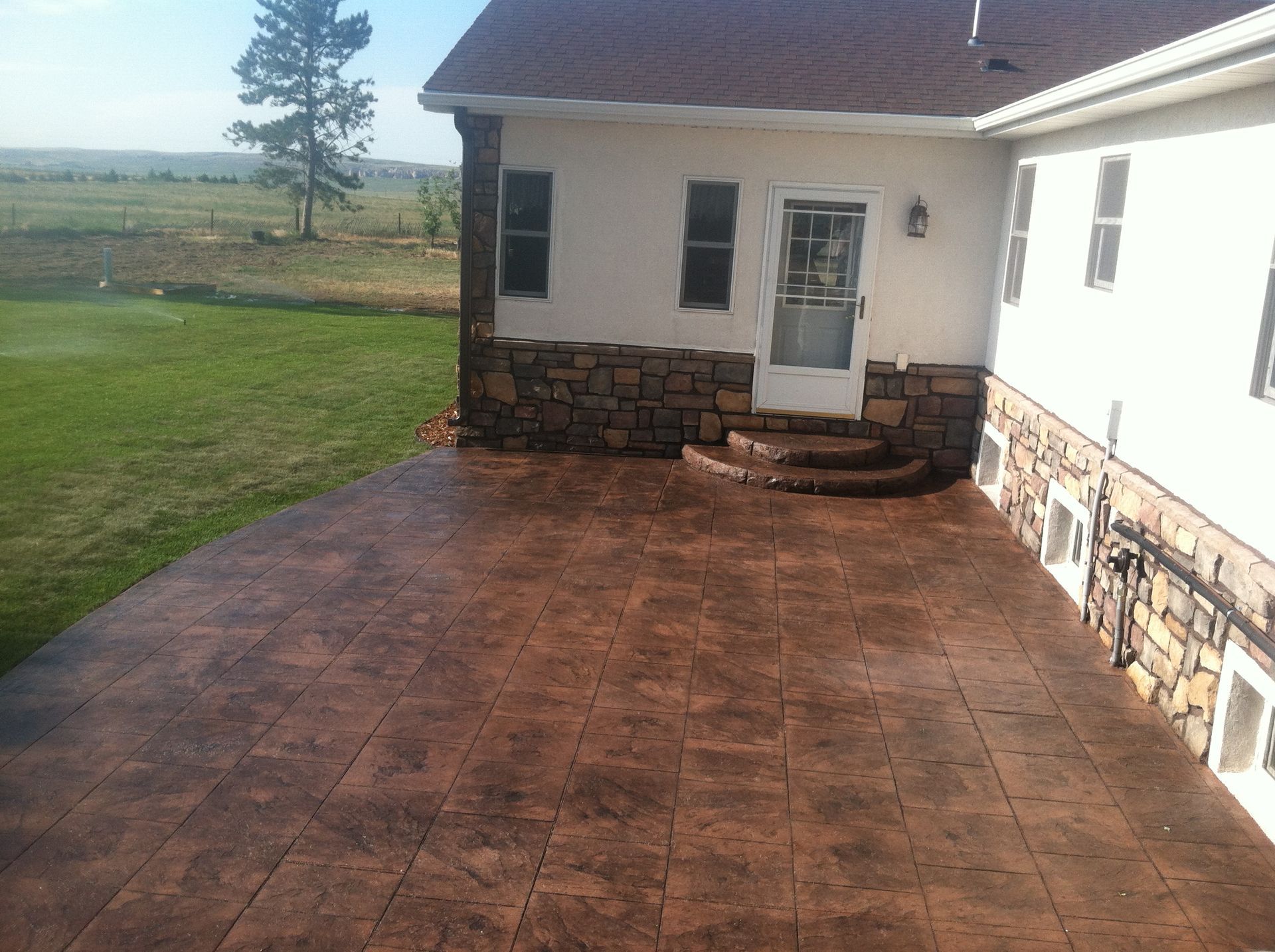 Brown stamped concrete patio with stone accents, leading to a house with a door and windows, surrounded by grass and a field.
