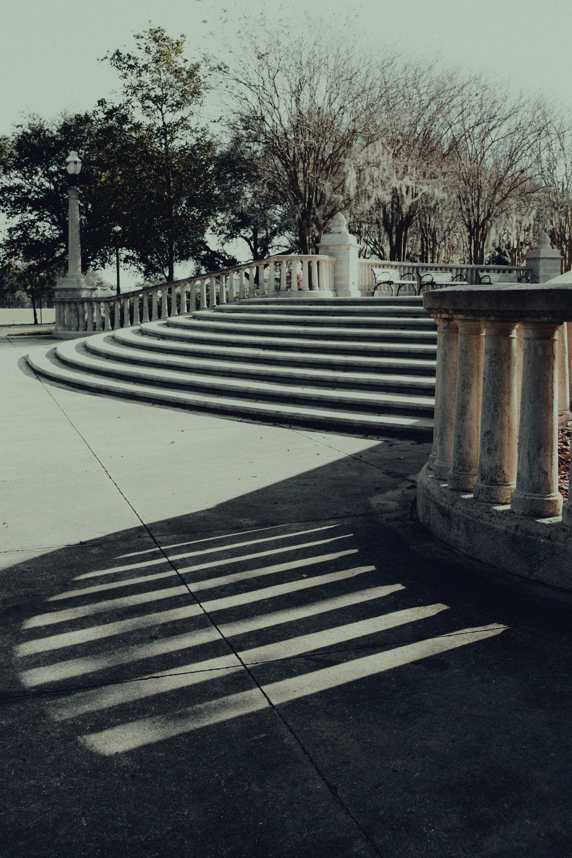 Curved stone steps lead to a park area with a balustrade casting long, striped shadows across the pavement.