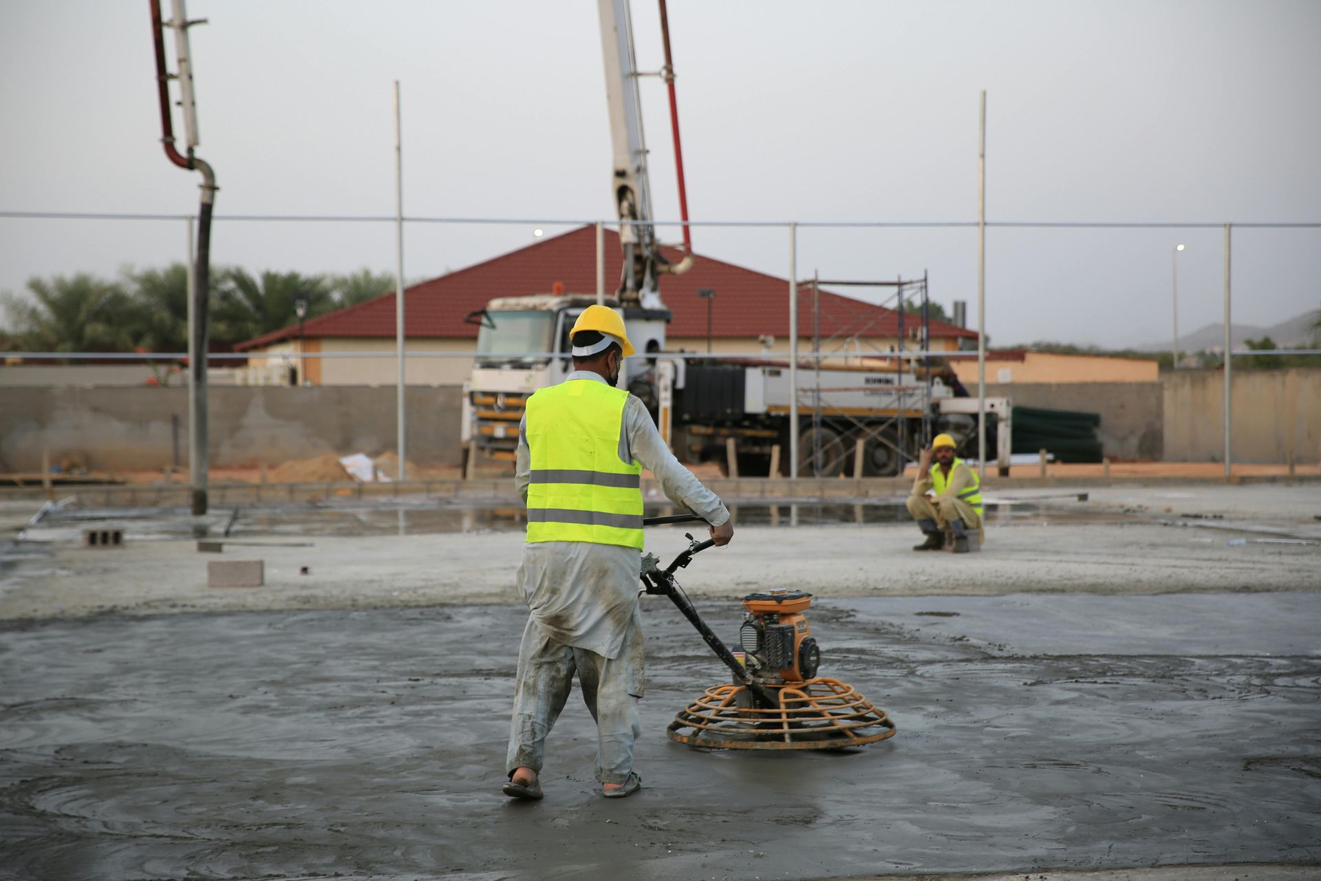 Construction workers smoothing wet concrete at an outdoor site, one in a neon vest in the foreground