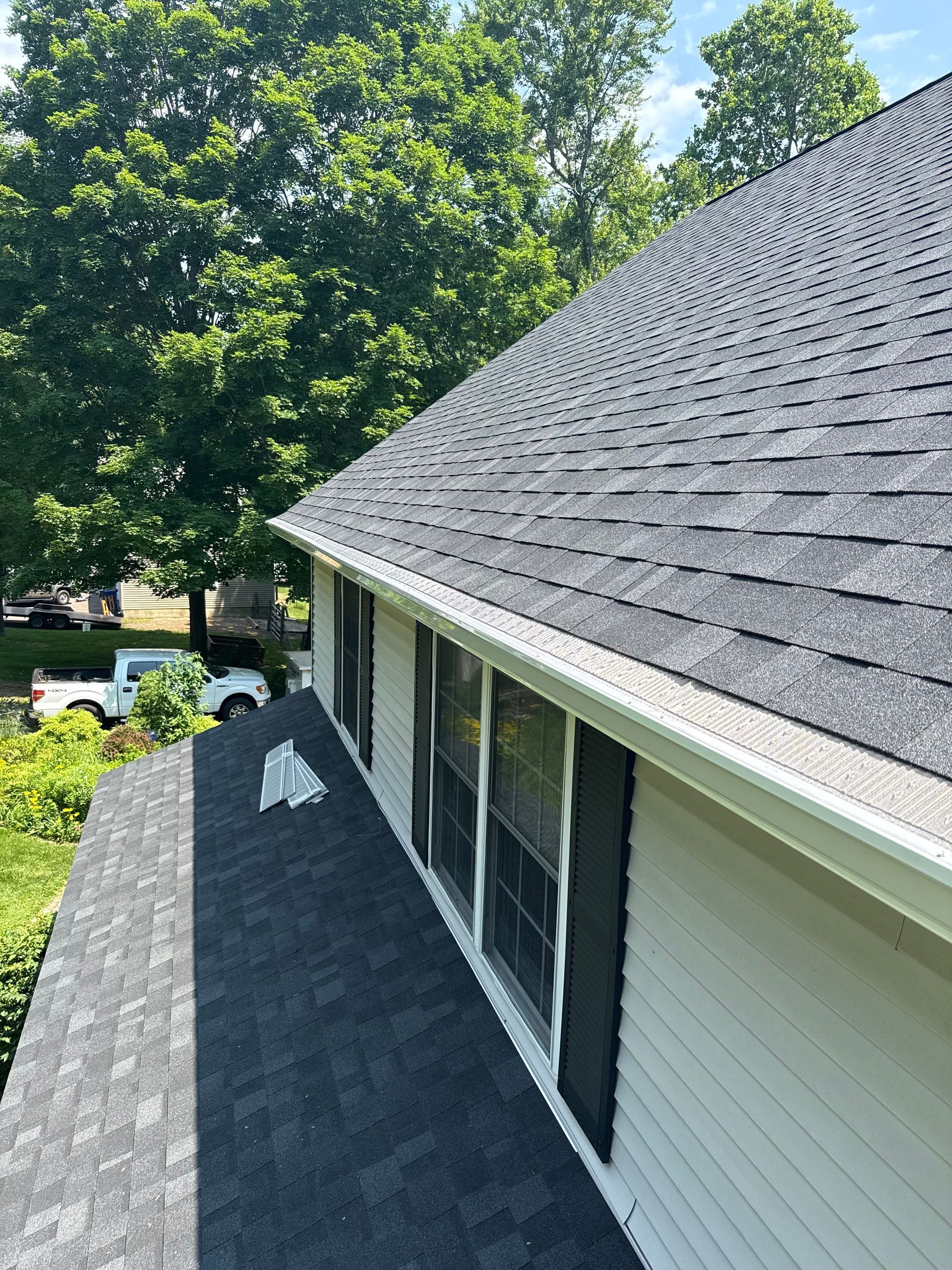 A house roof covered in gray shingles, with white trim and gutters, and lush green trees in the background.