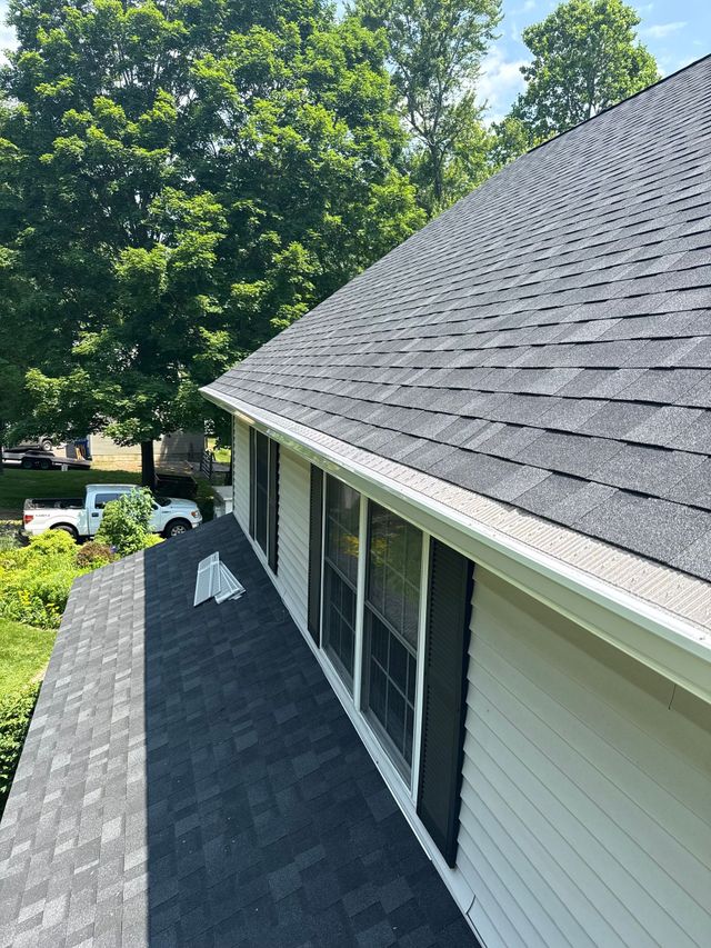 A house roof covered in gray shingles, with white trim and gutters, and lush green trees in the background.