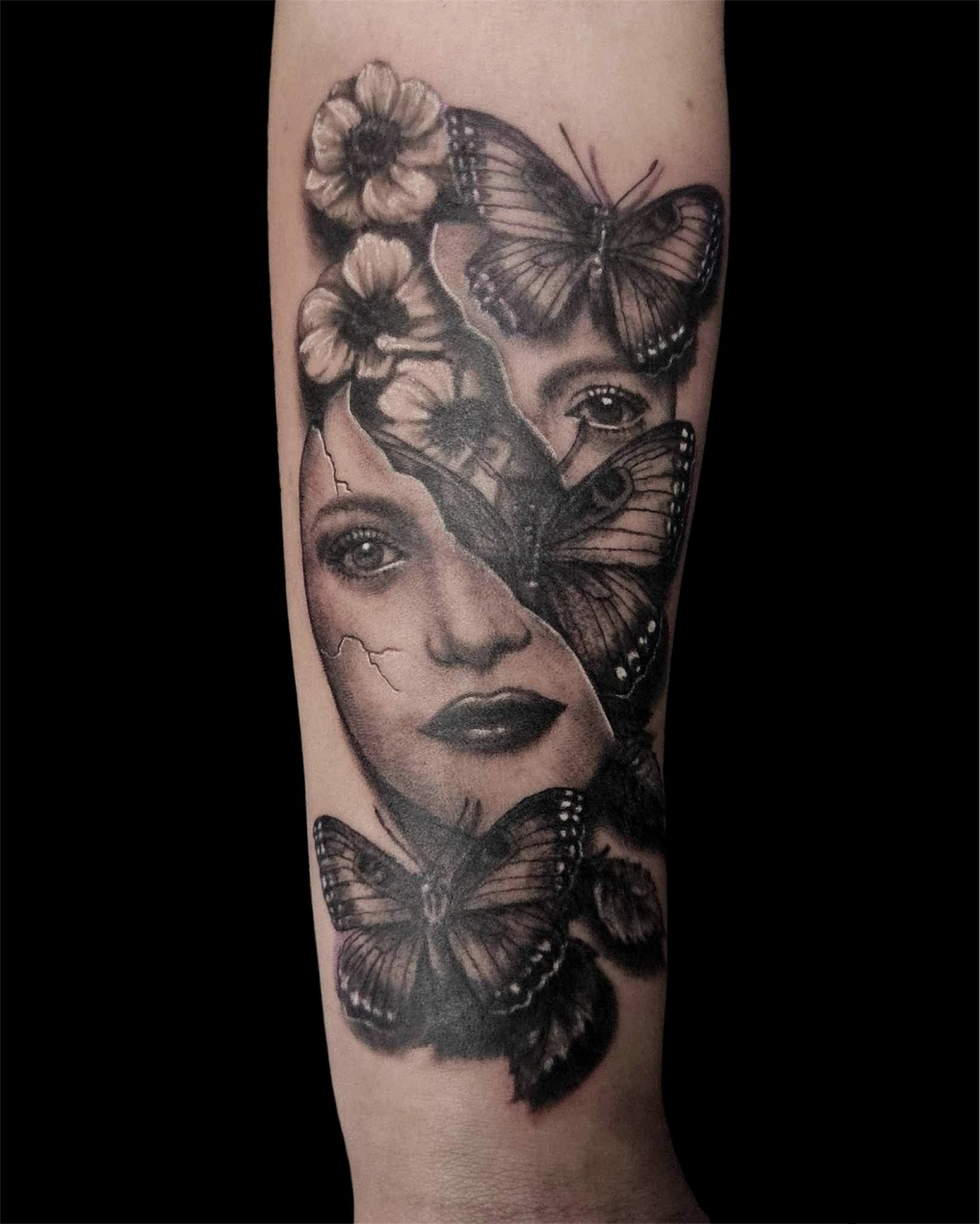 Black-and-gray tattoo of a woman’s face with butterflies and flowers on a forearm
