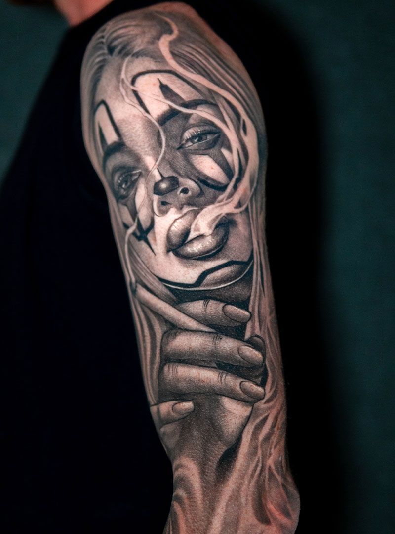 Black-and-gray tattoo of a smiling clown face on a forearm, with a hand gripping the lower face