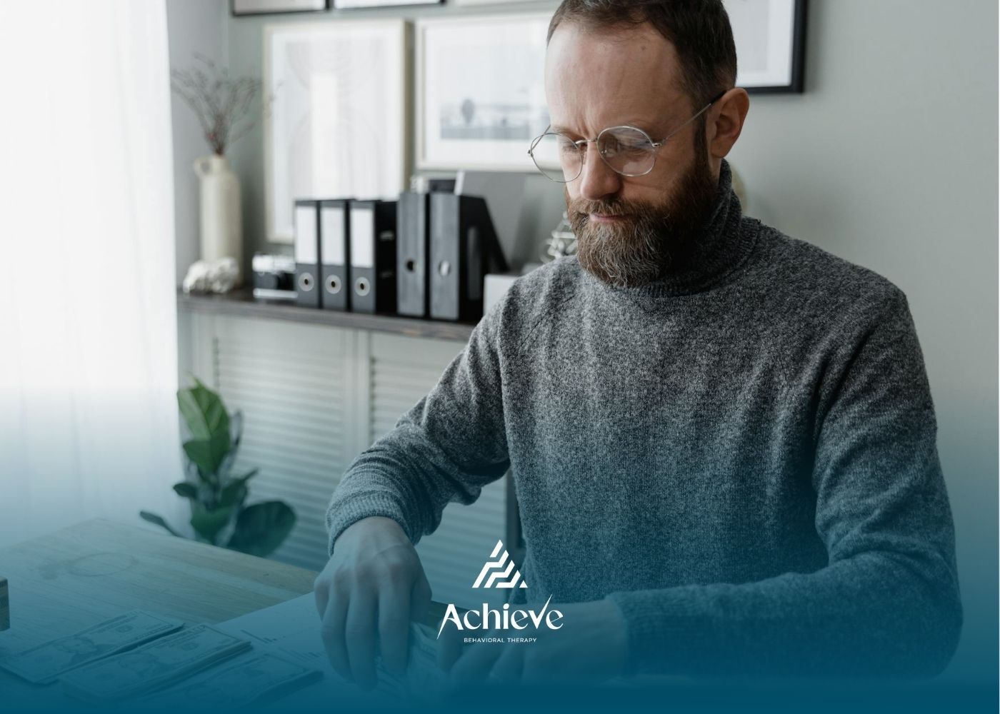 An RBT in gray sweater counts stacks of U.S. dollar bills at a desk in modern ABA therapy office.