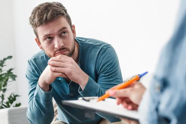 A man is sitting on a couch talking to a person holding a clipboard.