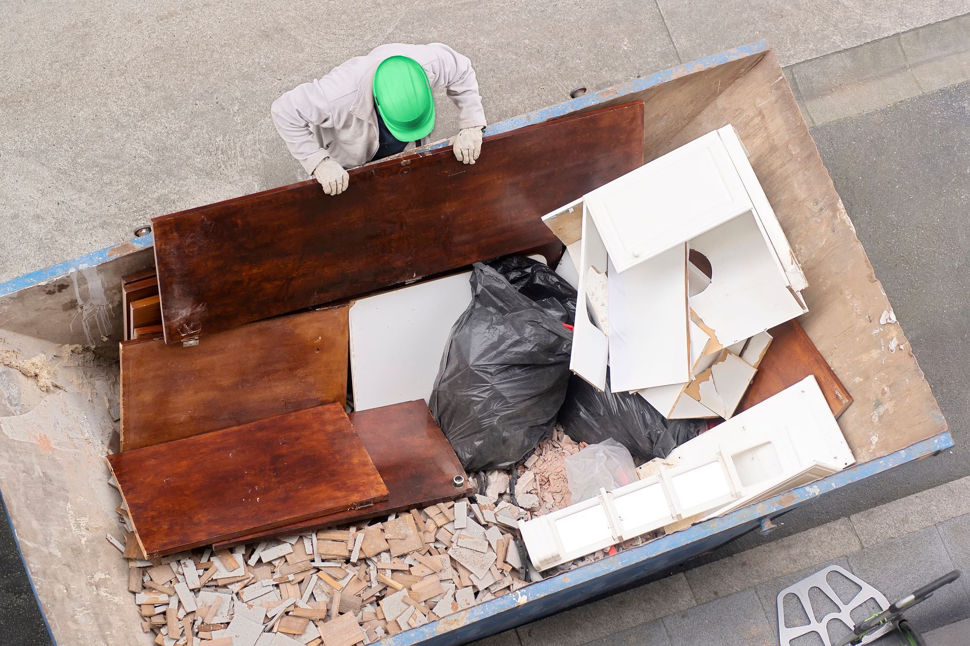 Person in green hard hat stands over dumpster filled with trash and wood debris.