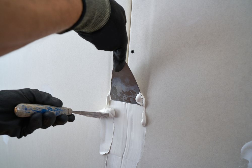 Person applying white joint compound to a wall seam with a putty knife, wearing black gloves.