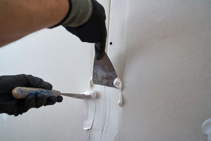 Person applying white joint compound to a wall seam with a putty knife, wearing black gloves.
