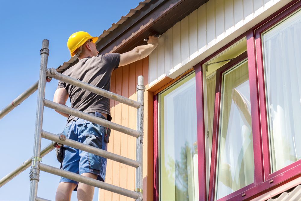 Painter on a ladder painting the trim of a house with a yellow hard hat on a sunny day.