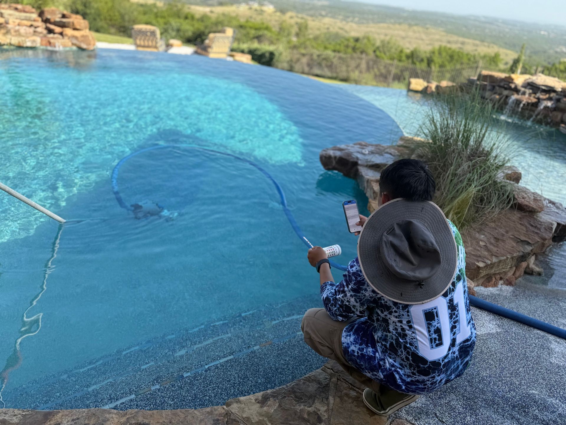 Person cleaning a blue infinity pool with a long-handled brush, hat on, overlooking a scenic landscape.